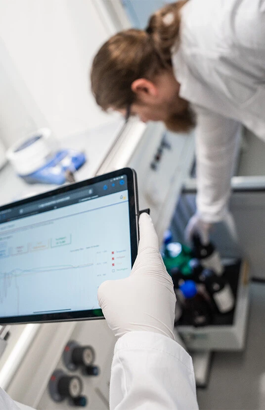 Ein Laborbild mit einer Person, die ein Tablet hält, um Daten anzuzeigen, während eine zweite Person im Hintergrund mit Flaschen arbeitet.